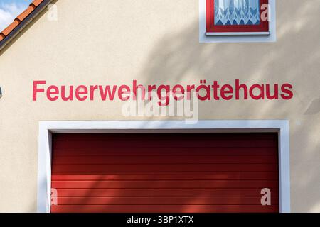 Close-up of a building with "Feuerwehrgerätehaus" in red lettering above a closed red garage door and a window with a white curtain. Stock Photo