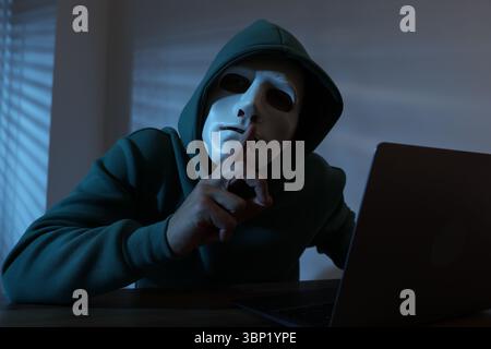 Anonymity. Masked man with laptop showing hush gesture at table in dark Stock Photo