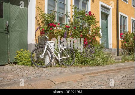 White bicycle parked by a yellow house with blooming flowers on cobblestone Adelgatan in Lund, Sweden, a charming summer street, June 30, 2025 Stock Photo
