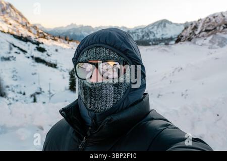 A man wearing a black jacket and a knit scarf is standing in the snow. He is wearing glasses and a hat Stock Photo