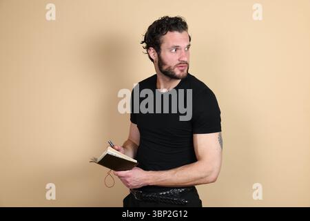A young man in a black t-shirt and pants holds a notebook and pen, looking back over his shoulder with a surprised or alert expression. Tattoo visible Stock Photo