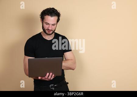 A young man in a black t-shirt and pants is shown in side profile, looking down and focused while working on a laptop he is holding. Studio portrait o Stock Photo