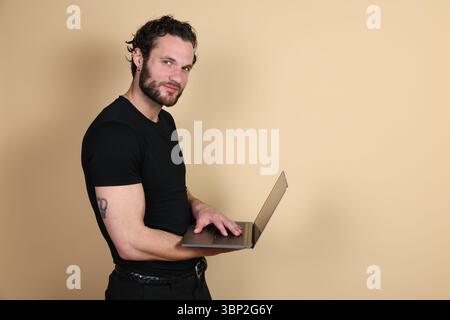 A confident and friendly man with a beard smiles at the camera while holding a laptop, standing against a plain beige studio background with copy spac Stock Photo