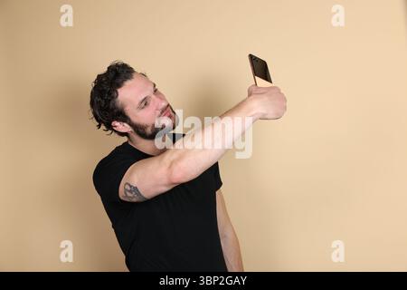 A young man with a visible arm tattoo focuses on his smartphone screen while taking a selfie, against a plain beige studio background. Stock Photo