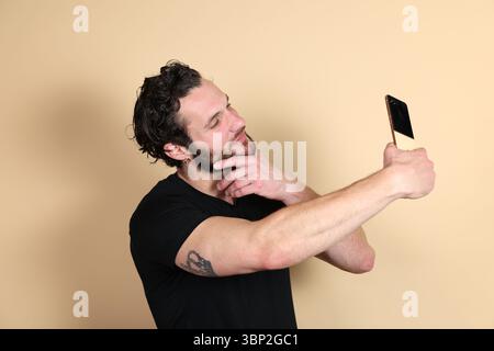 A man with a tattoo thoughtfully holds his chin while taking a selfie with his smartphone, looking at the screen from a slight angle, against a plain Stock Photo
