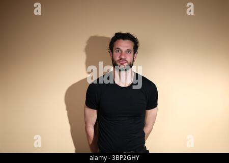 A young bearded man in a black t-shirt stands calmly facing the camera with a neutral expression, his hands clasped behind his back, against a plain s Stock Photo