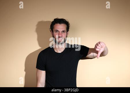 A bearded man in a black t-shirt looks directly at the camera with a serious expression while gesturing thumbs down, clearly indicating disapproval or Stock Photo