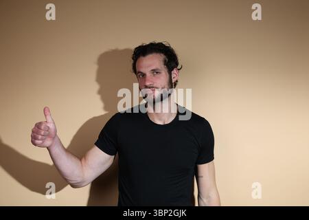 A confident and smiling bearded man in a black t-shirt gives a thumbs-up gesture directly to the camera, expressing clear approval and positivity agai Stock Photo