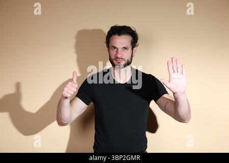 A young man looks at the camera making a dual gesture: a thumbs up with one hand for approval, and a stop sign with the other palm forward, indicating Stock Photo