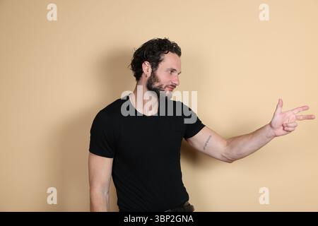 A young man in a black t-shirt makes a three-finger gun gesture, pointing and looking to the side with a playful or focused expression. Studio shot on Stock Photo