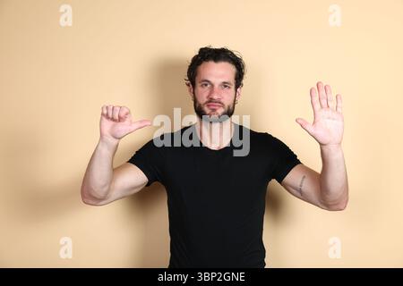 A young man looks at the camera making a dual gesture: a thumbs up angled towards himself (self-approval/I'm good) with one hand, and a stop sign with Stock Photo