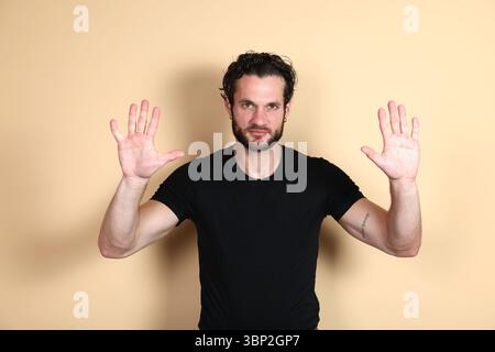 A young man in a black t-shirt looks directly at the camera with both hands raised, palms open and facing forward, conveying a stop, surrender, "hands Stock Photo