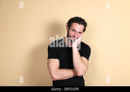 A young man in a black t-shirt looks directly at the camera with a bored, tired, and fed-up expression, resting his cheek on his hand, arm crossed. St Stock Photo