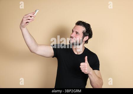 A young man in a black t-shirt smiles while taking a high-angle selfie with his smartphone, arm extended upwards, and giving a thumbs-up. Studio shot Stock Photo