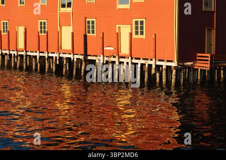 Picturesque red Scandinavian fishing houses on wooden stilts reflecting in the calm harbor water during a beautiful golden hour. Stock Photo