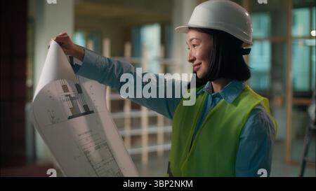 Woman civil engineer in hard hat Asian girl inspector looking at building plan construction paper draft electricity Chinese Korean Japanese female Stock Photo