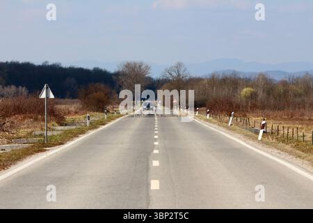 A straight, two-lane rural road stretches into the distance, bordered by dry grass and shrubs, with a single car approaching and white roadside marker Stock Photo