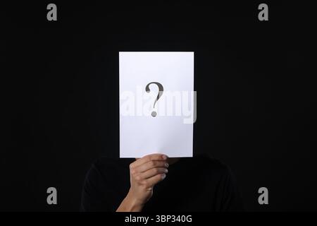 Man hiding behind paper sheet with question mark on dark background Stock Photo