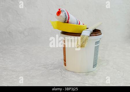 A close-up image of a white plastic paint bucket with a brown lid, topped with a paint roller, masking tape, and a paintbrush. The items are arranged Stock Photo