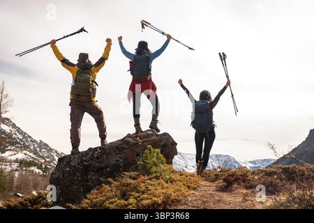 Happy tourists stand on a cliff, admiring the breathtaking view of the valley and mountain peaks. Joy and delight from reaching the goal in hike. Raise their hands up with trekking poles Stock Photo