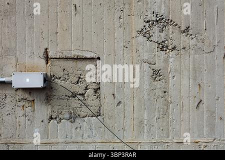 Weathered concrete wall features rectangular patch with exposed rebar and electrical box with visible wire. Its rough surface adds industrial feel. Stock Photo
