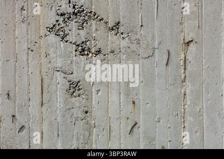 Concrete wall displays signs of wear and erosion in exposed aggregate and protruding rusty nails. Gray hues highlight rough texture's imperfections. Stock Photo