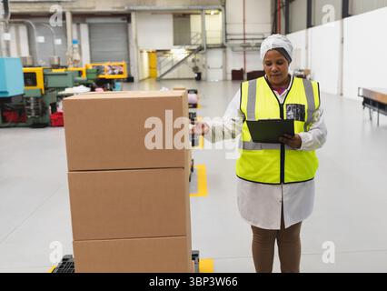 Woman inspecting stacked cardboard boxes on pallet in warehouse while holding clipboard, copy space Stock Photo