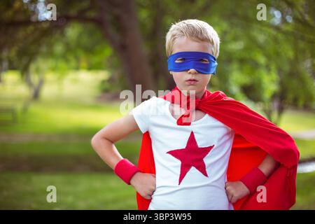 Child standing confidently in grassy park wearing red cape and wristbands with blue eye mask Stock Photo