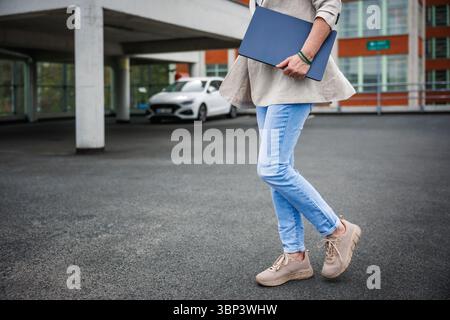 Woman entrepreneur crossing parking lot with laptop in hand, casual street business fashion Stock Photo