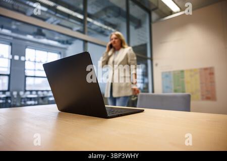 Open laptop standing on office desk while businesswoman talking on mobile phone in background. Leadership in corporate business Stock Photo