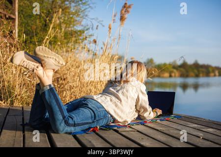 Woman lying on pier at lake, working remotely on laptop during sunny spring day. Freelance work Stock Photo