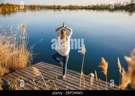 Woman practicing yoga exercise and tree pose on pier at lake. Fitness active lifestyle for mental wellbeing Stock Photo