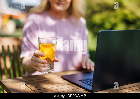 Woman drinking water with slice of orange while working on laptop in outdoor cafe Stock Photo