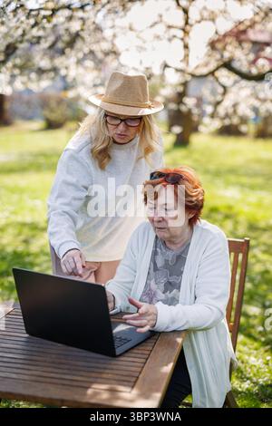 Senior woman looking at laptop with interest while her adult daughter is helping how to use it. Using laptop in spring garden Stock Photo
