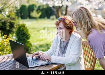 Senior mother with her adult daughter are using laptop in garden. Multi-generation women watching computer screen outdoors Stock Photo