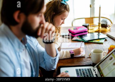 Girl doing homework while sitting next to father working on laptop at home Stock Photo