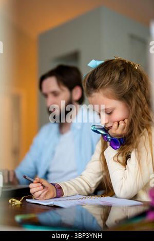 Girl with blond hair studying while sitting next to father working at home Stock Photo