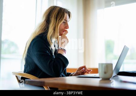 Thoughtful female freelancer working on laptop while sitting on chair at home office Stock Photo