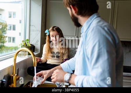 Girl sitting on counter and talking with father washing dishes in kitchen at home Stock Photo