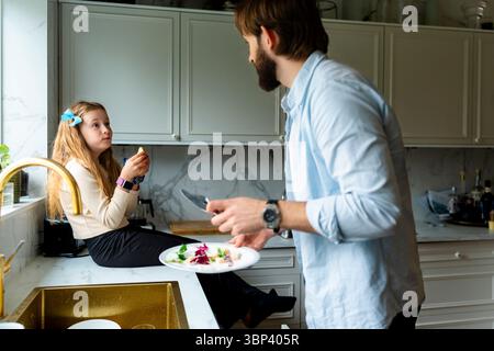 Girl eating fruit while talking with father working in kitchen at home Stock Photo