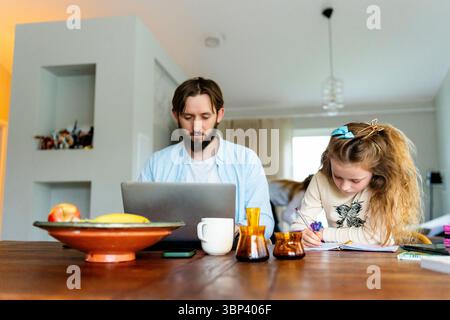 Father working on laptop while sitting next to daughter doing homework Stock Photo