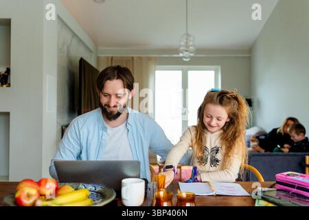 Smiling father working on laptop next to daughter studying while sitting at home Stock Photo