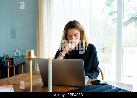 Thoughtful freelancer with hand on chin working on laptop at home office Stock Photo