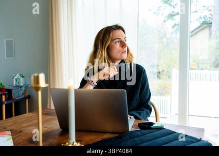 Female freelance worker looking away while sitting with laptop on table at home office Stock Photo