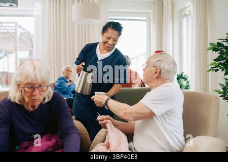Smiling caregiver serving tea to elderly man sitting in retirement home Stock Photo