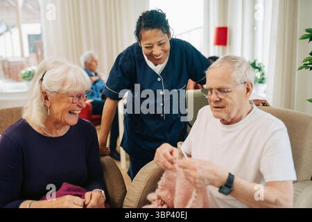Happy female healthcare worker and senior woman looking at elderly man knitting wool at nursing home Stock Photo