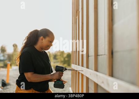 Concentrated female carpenter drilling nail on wooden plank outside house Stock Photo