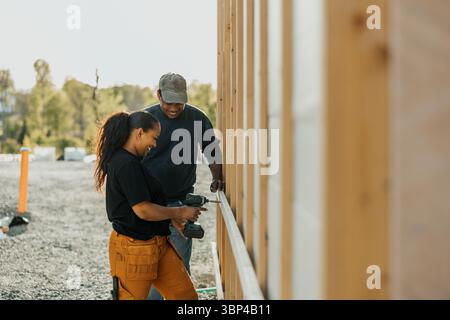 Female worker using drill on wooden plank standing next to colleague outside house Stock Photo