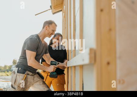 Man drilling wooden plank outside house with help of woman Stock Photo