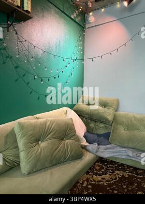 The photo shows a corner interior with a soft green cushioned sofa, several backrest pillows, and some loosely placed blankets. The wall is painted in Stock Photo